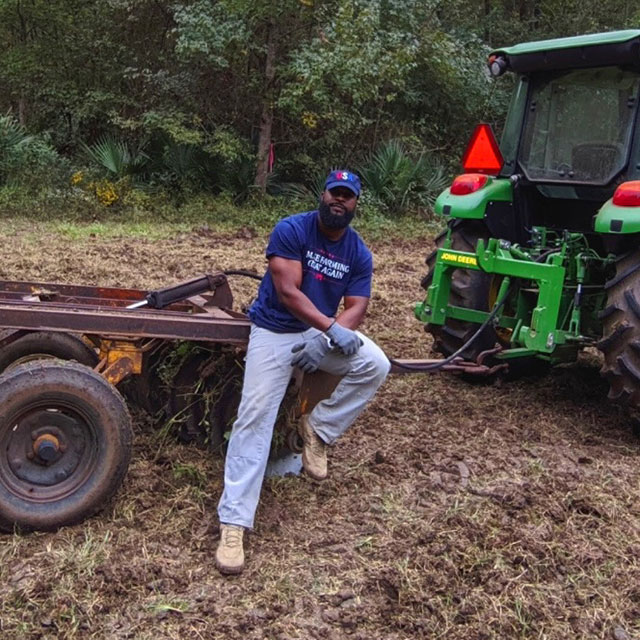 Christopher Joe, AL, wearing a blue t-shirt that says “Maker Farming Great Again” and light blue jeans leaning on tractor equipment.